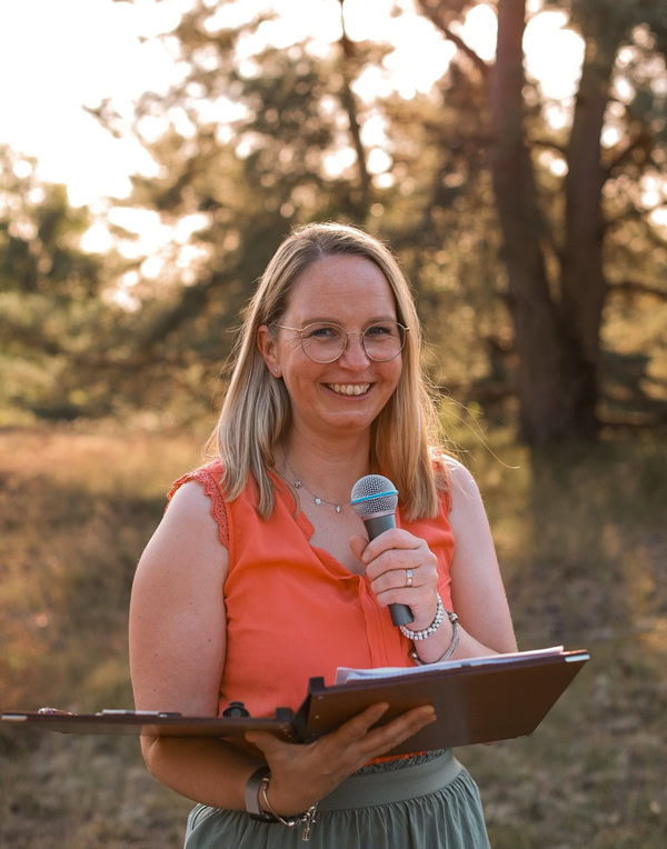 Die Freie Rednerin Meike steht in der Natur im Sonnenlicht, hält ihr Mikro und ihre Ledermappe in der Hand.