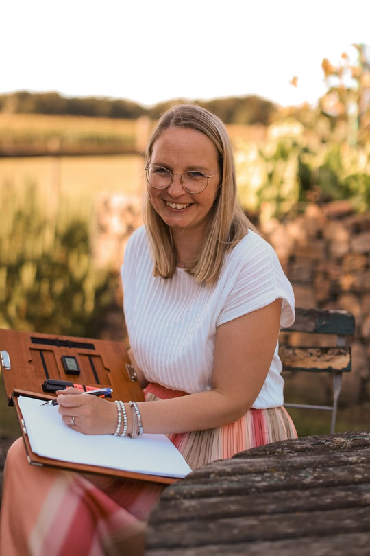 Die freie Rednerin Meike sitzt vor einem sonnigen Feld und schreibt in ihre Mappe.