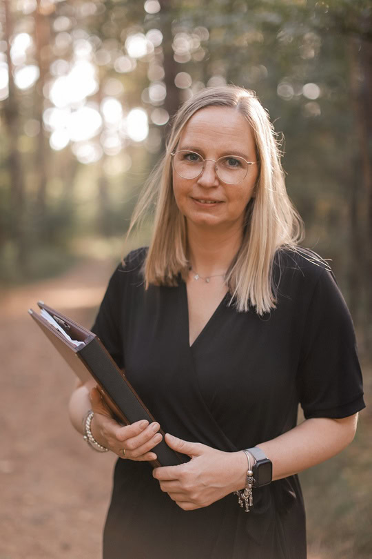 Trauerfeier in der Region Hannover. Meike hält ihre Rednermappe in der Hand und steht in einem Wald.