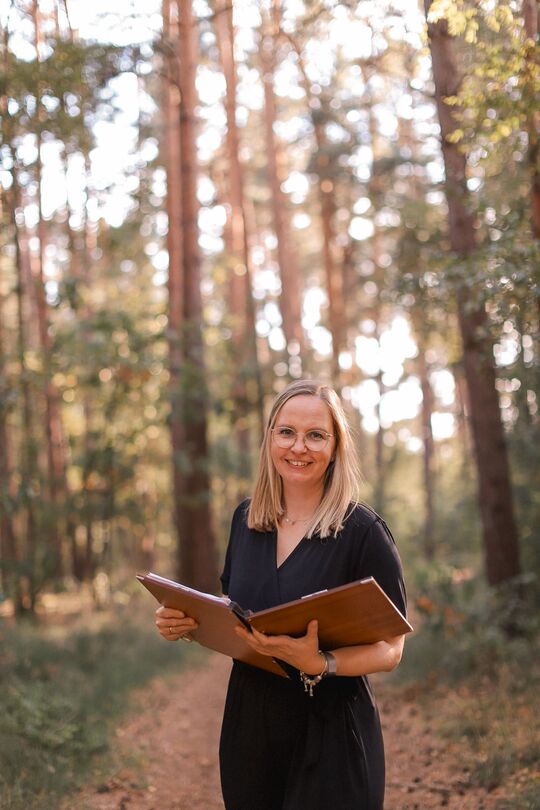 Freie Rednerin in der Region Hannover. Meike steht im Wald, schaut freundlich in die Kamera und hält ihre braune Ledermappe in der Hand.